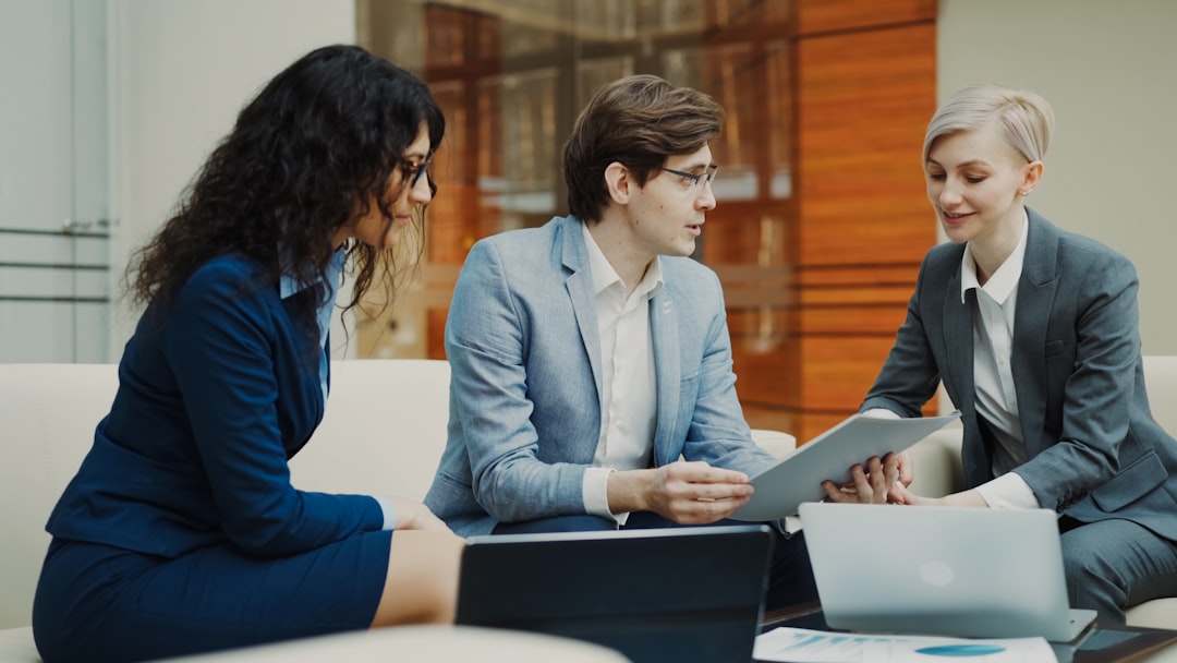 Three business professionals in a collaborative meeting discussing strategy, seated on a modern office sofa with documents and laptops